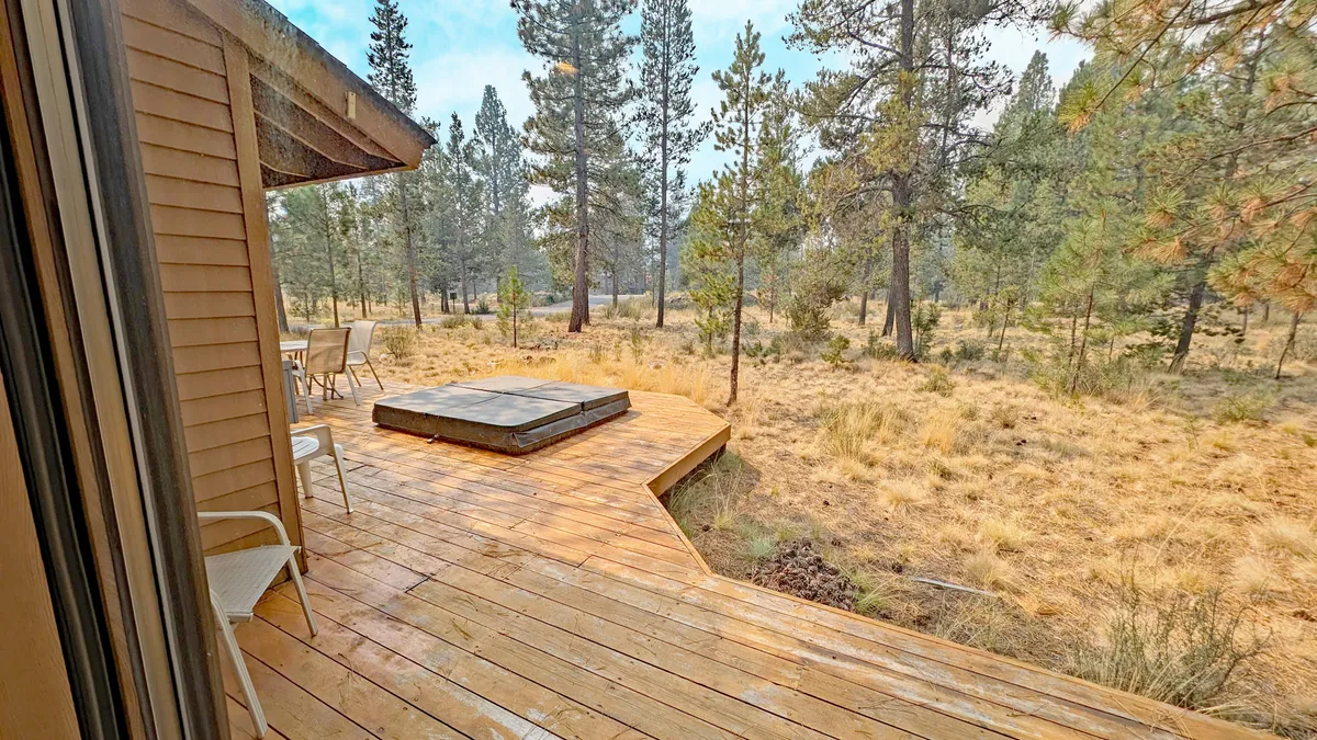 A panoramic exterior view of the deck with the hot tub in the foreground, outdoor dining area, and surrounding natural landscape. The expansive setting captures both relaxation and the beauty of the outdoors, ideal for stargazing or morning coffee.