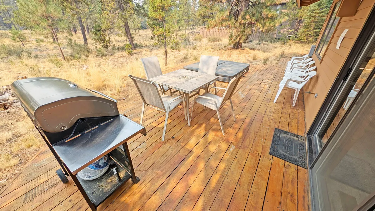 The image features a grill, outdoor dining table, and comfortable seating set on a spacious deck under the shade of tall pine trees. A sense of community and enjoyment is conveyed, perfect for gatherings or quiet meals outdoors.
