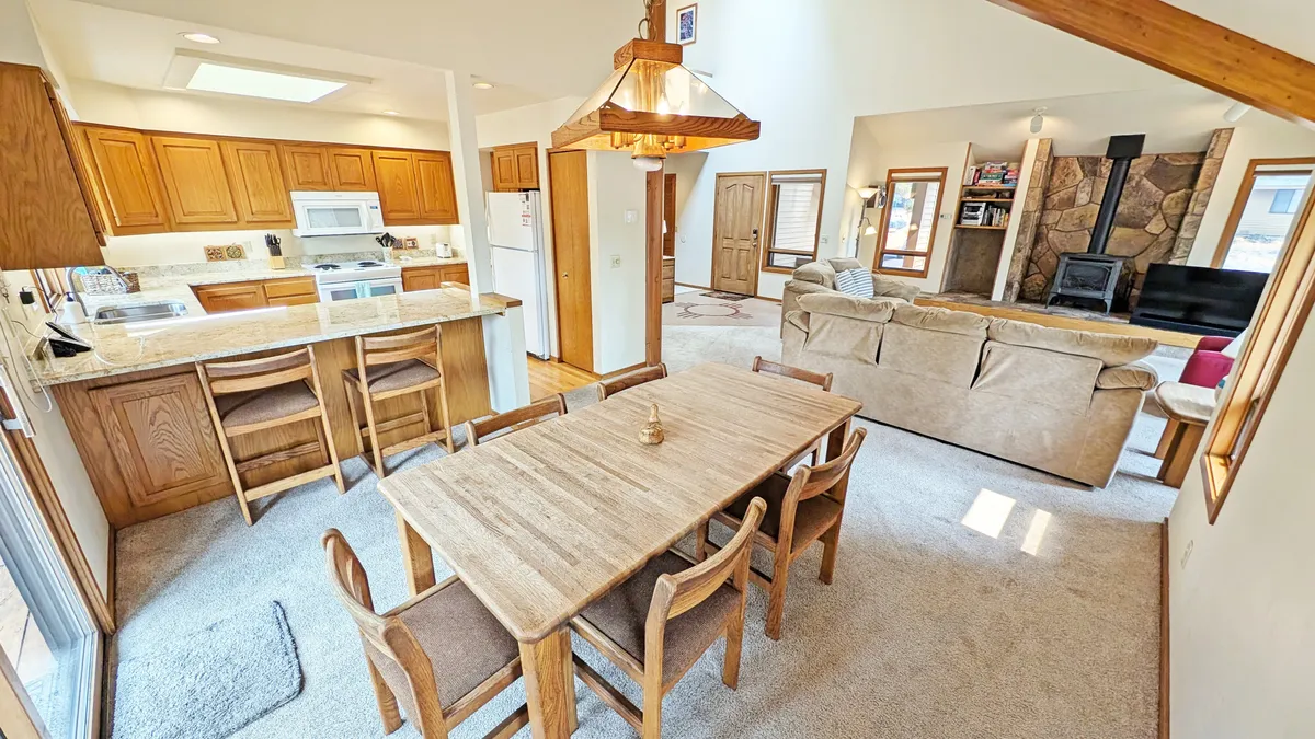 A wide view of the open-concept living, dining, and kitchen area. The scene features a cozy sofa set near a fireplace, a spacious dining table ready for family meals, and a kitchen island that connects all three spaces. The area is well-lit with natural light and arranged for both comfort and social interaction.