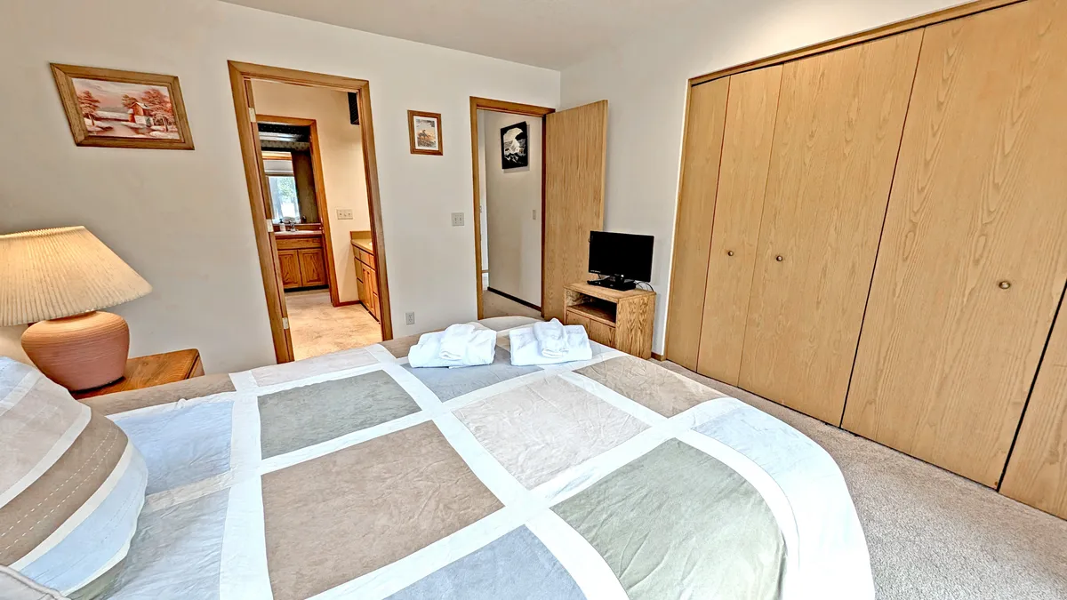 A light-filled bedroom with a queen bed draped in a quilt, a bedside lamp, closet doors, and a television. Framed artwork and soft natural light create a comfortable, welcoming atmosphere ideal for relaxation and privacy.