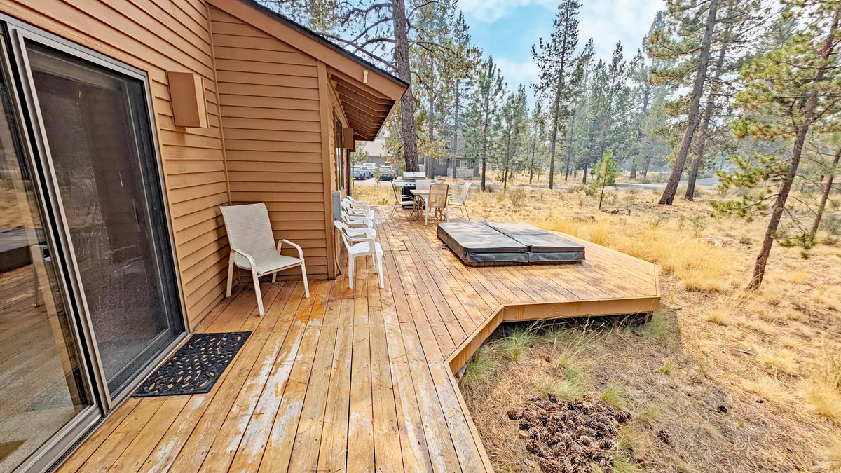 A spacious wooden deck nestled among towering trees, featuring a bubbling hot tub, lawn chairs, and ample outdoor seating. The setting is tranquil, with lush greenery surrounding the deck and soft, natural light creating a relaxing ambiance.