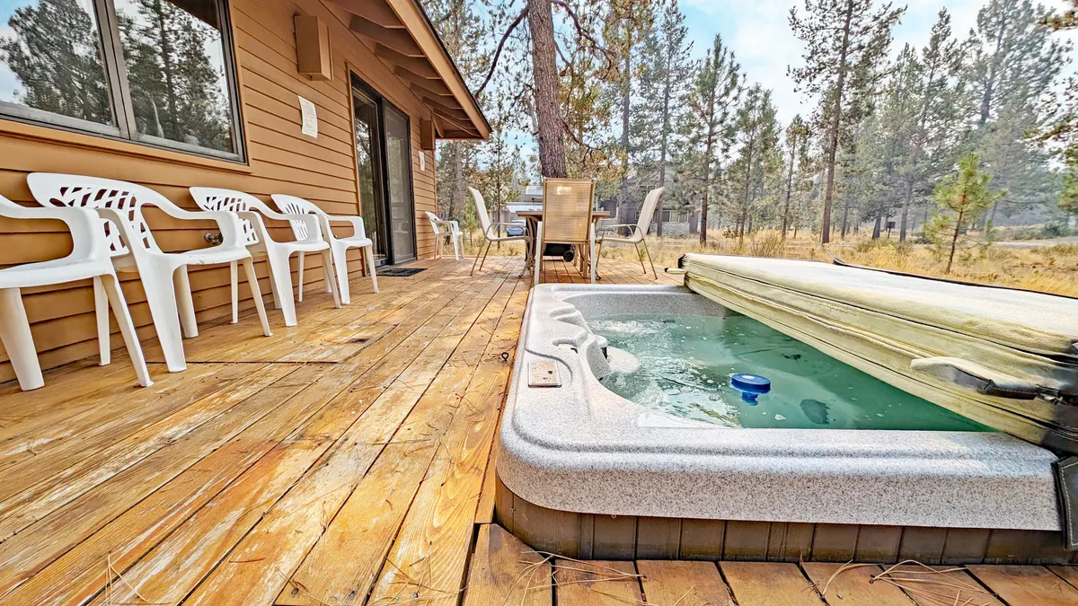 An inviting shot of a hot tub and spacious wooden deck with a seating area, embraced by greenery and open sky. The setting hints at outdoor leisure, conversation, and enjoyment after a day of activities.