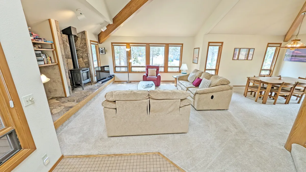 A comfortable living room scene with a stone fireplace as the focal point, large windows letting in natural light, a plush sofa, and a dining table nearby. The setting is styled for relaxation and social occasions, evoking warmth and togetherness.