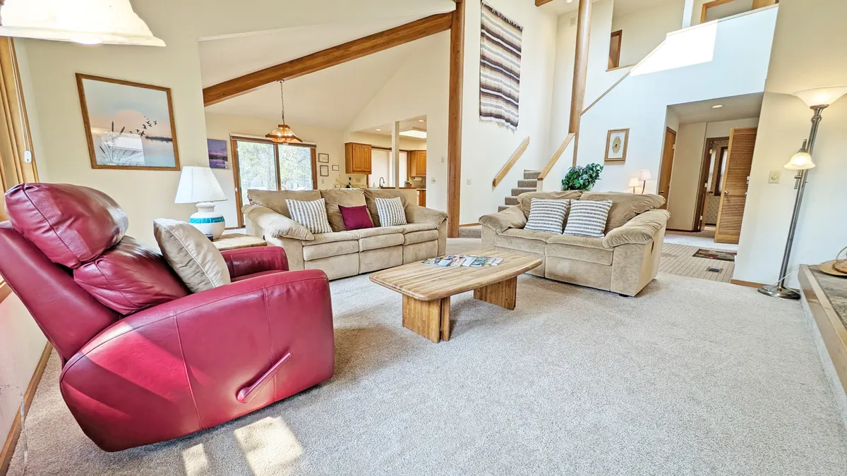 The image features a warm, inviting living room with a plush red recliner, comfortable beige sofas arranged around a wooden coffee table, and expansive windows that flood the space with natural light. The room's layout encourages gathering and relaxation, ideal for family and friends to unwind together after a day of adventure in Sunriver.