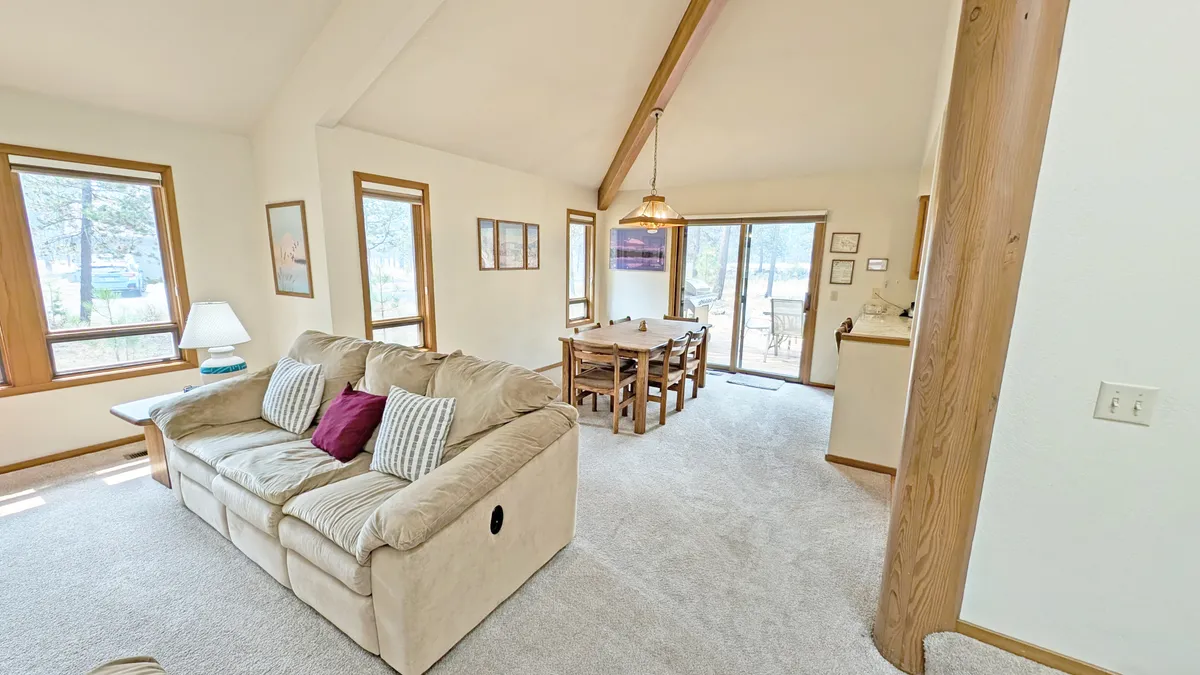 This image features a cozy living space with a sofa, dining table, and large windows that allow natural light to pour in. The setup is ideal for group gatherings, shared meals, and relaxed conversations, with a strong connection to the outdoors through the visible windows.
